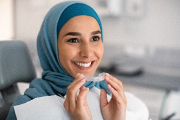 Woman smiling while holding Invisalign aligner at dental office