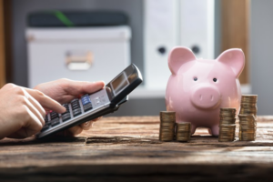 Person using a calculator with a piggy bank and coin stacks sitting nearby